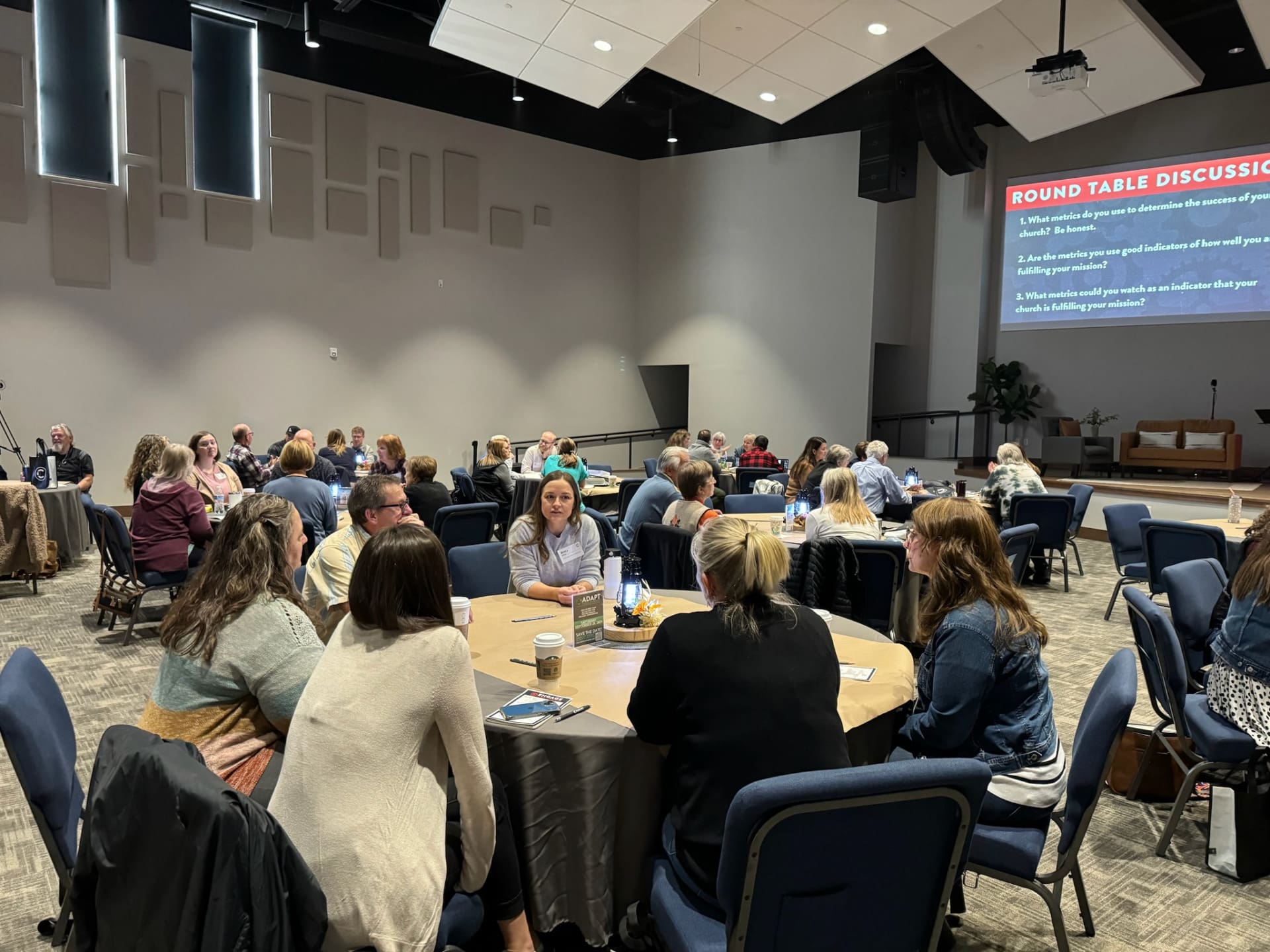 People sit in groups around round tables in a large room, engaged in discussion. A screen at the front displays “Round Table Discussion” with discussion questions focused on support for rural church leaders. The setting appears to be a workshop or seminar.