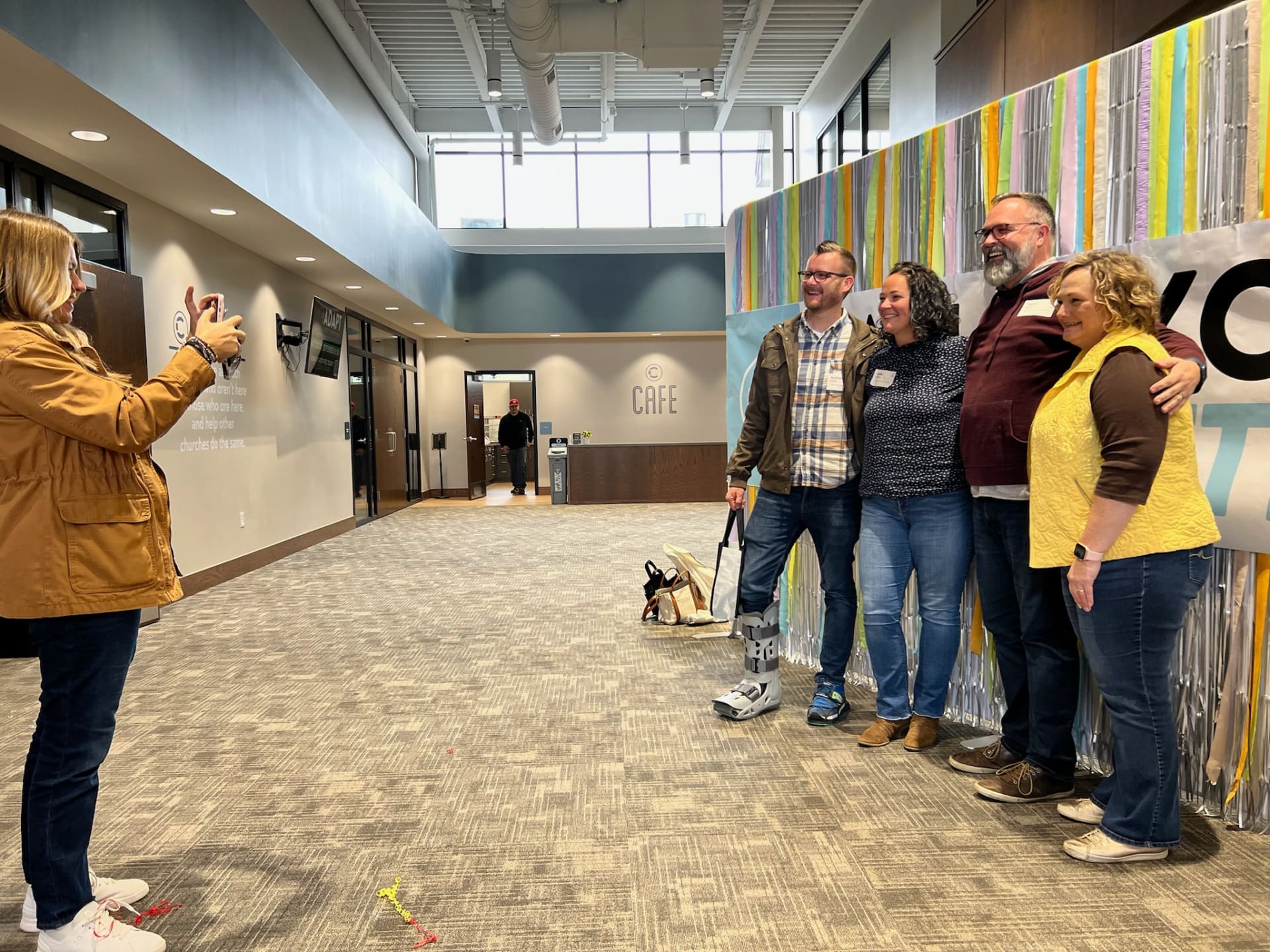 A woman takes a photo of five adults standing together and smiling in front of a colorful backdrop in a bright building lobby, capturing a joyful moment that highlights support for rural church leaders.