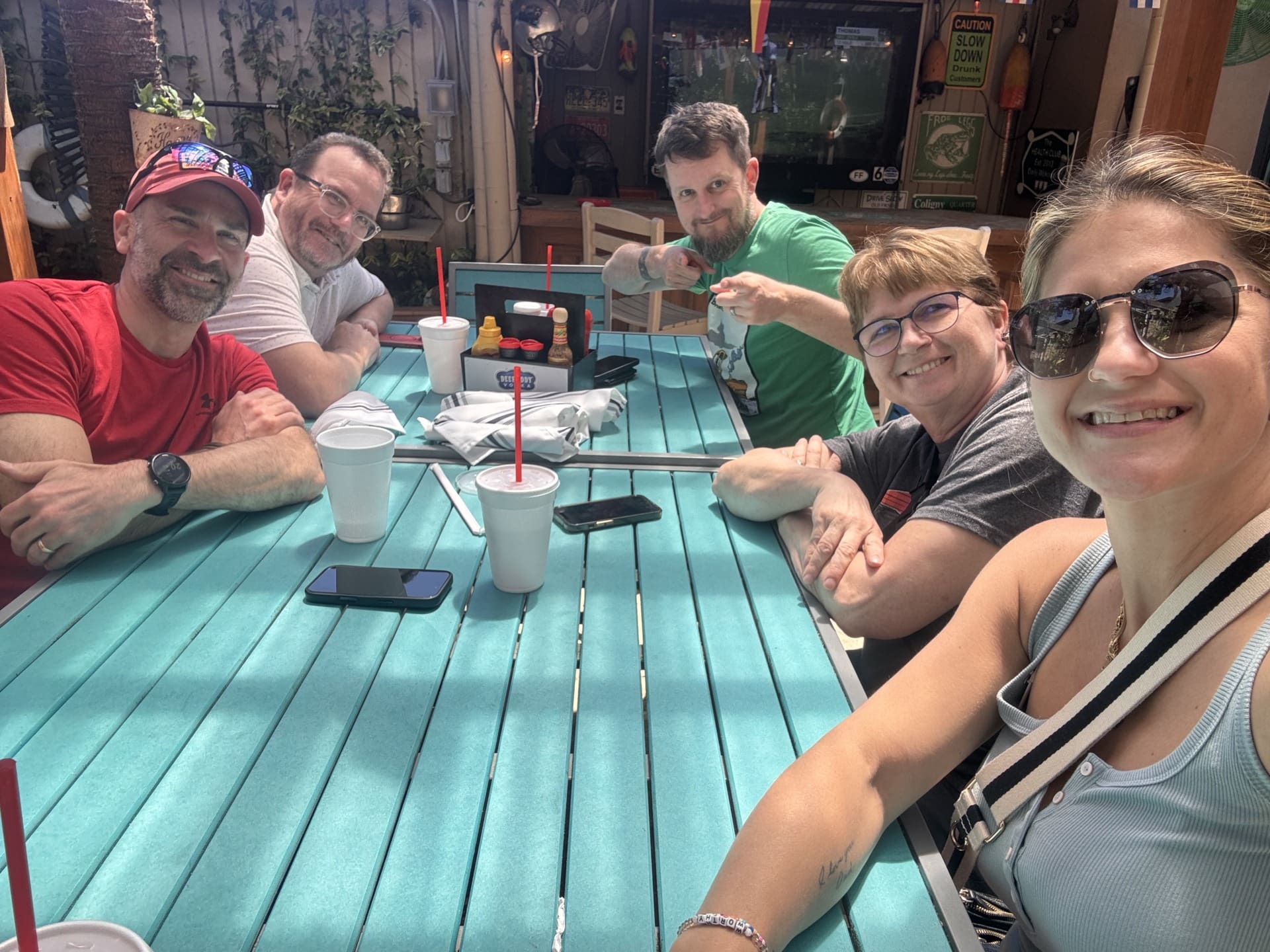 Five people pose for a selfie at a teal outdoor table at a restaurant. A woman in the foreground takes the photo, while others behind the table smile or point at the camera. Drinks and phones sit on the table.