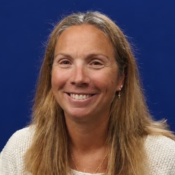 A woman with long light brown hair smiles at the camera. She is wearing a cream sweater, gold jewelry, and small cross earrings, with a solid blue background behind her—reflecting the warmth and dedication often seen in those providing support for rural church leaders.