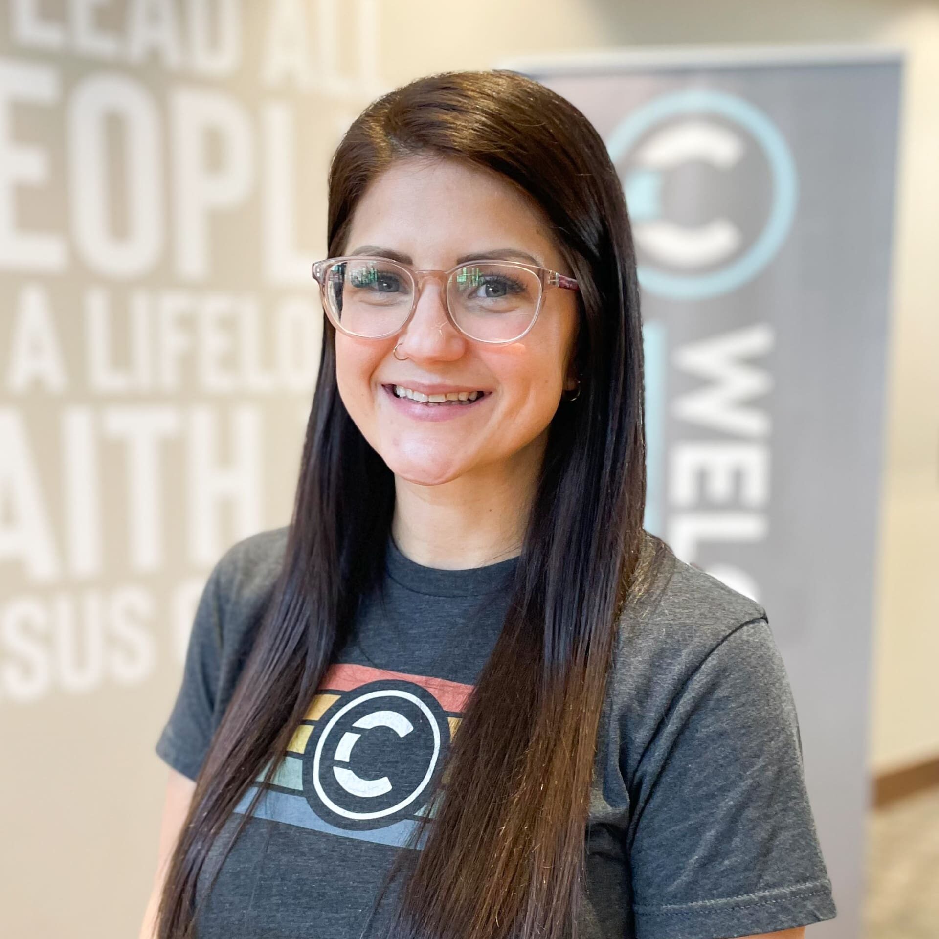 A woman with long dark hair and glasses smiles while wearing a T-shirt with a circular logo. There is a blurred banner and wall in the background with partially visible words.