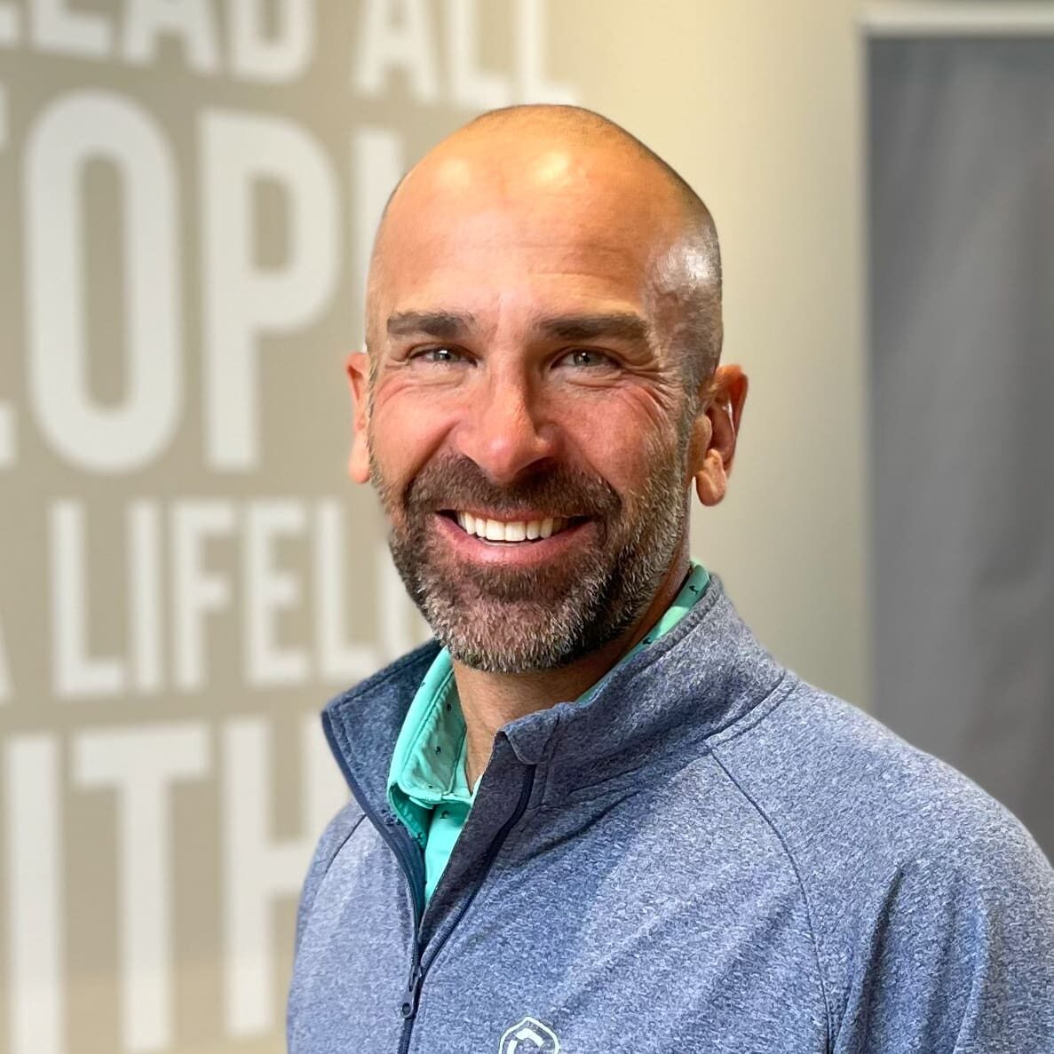 A smiling man with a bald head and beard, wearing a light blue zip-up jacket over a light green shirt, standing indoors with blurred text in the background.