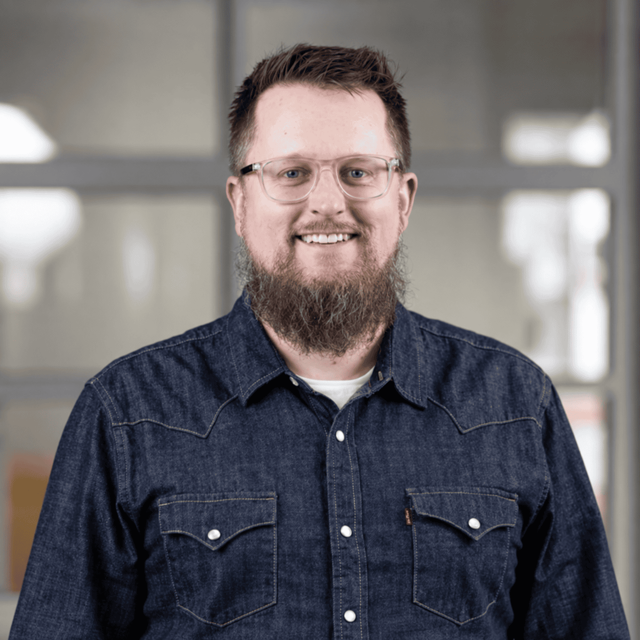 A man with glasses and a full beard, wearing a dark denim shirt, stands smiling in front of a blurred indoor background with large windows.