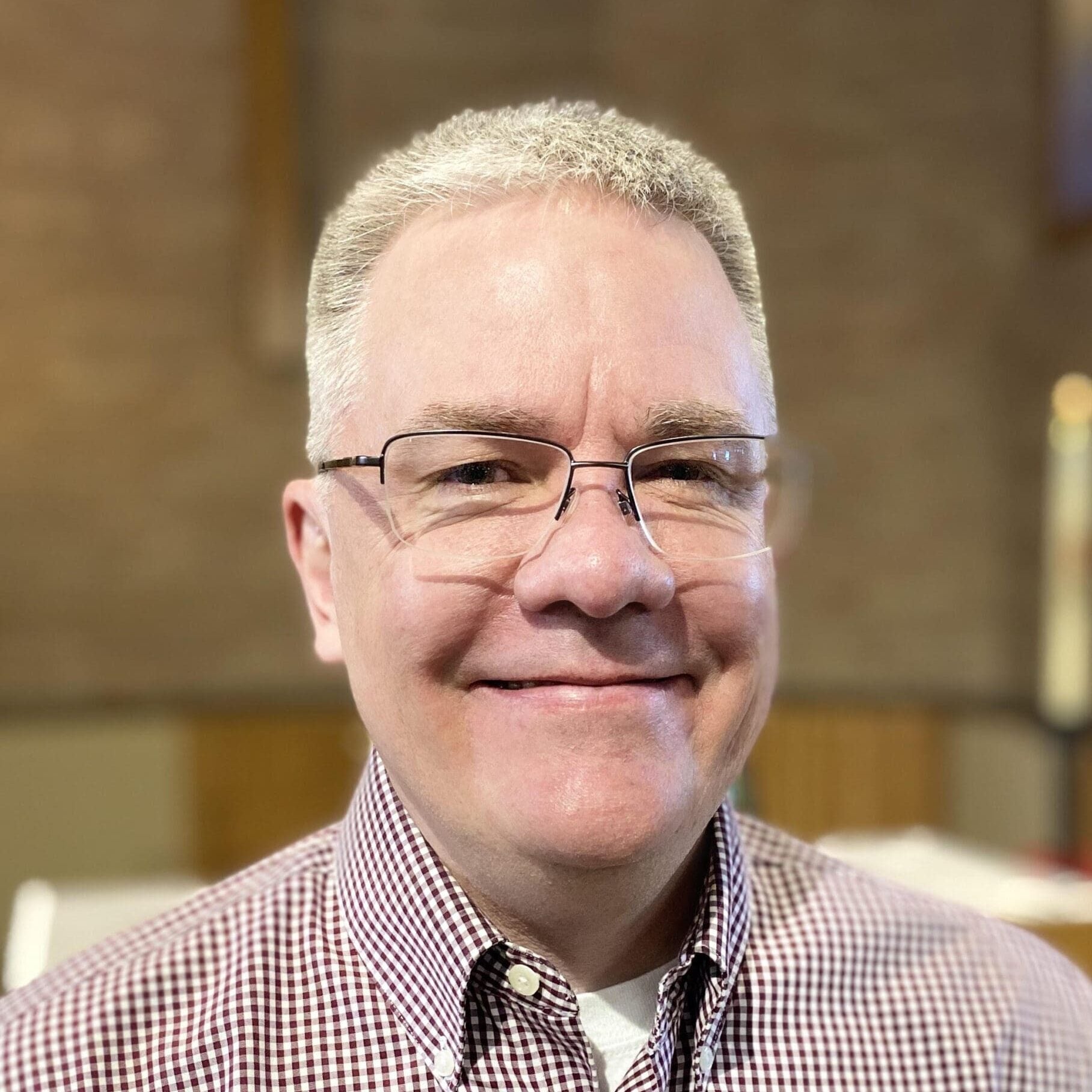 A middle-aged man with short gray hair and glasses smiles at the camera. He is wearing a checkered shirt and is standing indoors with a blurred background, embodying the warmth and dedication often seen in those providing support for rural church leaders.